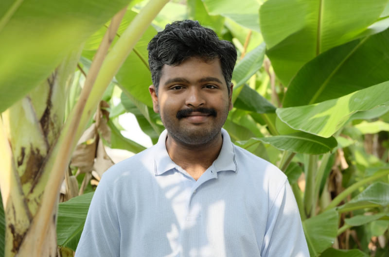 Siva in a light blue tee shirt, standing with a pleasant expression, surrounded by banana plants.
